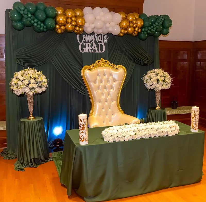 Green and gold graduation backdrop with a white throne chair and floral arrangements